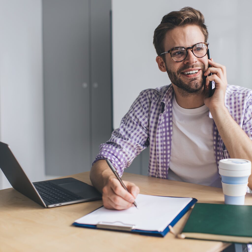 Man conducting customer complaint investigation talks on phone while writing on clipboard with notebook and laptop nearby
